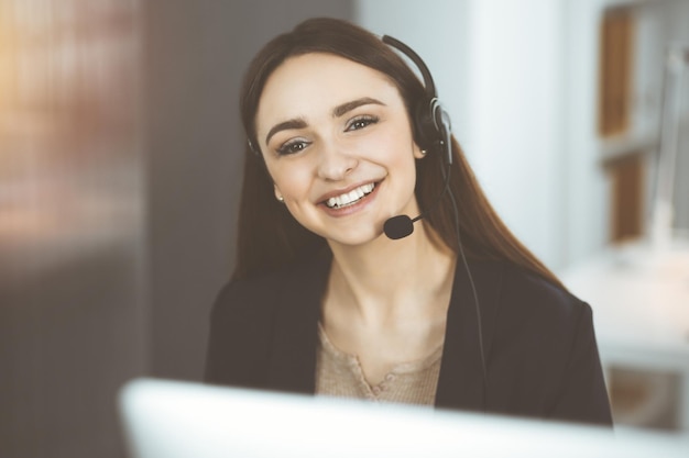 Młoda przyjazna dziewczyna w zestawach słuchawkowych rozmawia z klientem firmy, siedząc przy biurku w słonecznym biurze. Operatorzy call center w pracy.