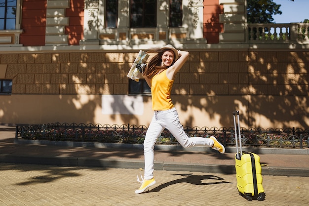 Młoda radosna podróżniczka turystyczna kobieta w żółtych letnich ubraniach casual, kapelusz z walizką mapę miasta, skoki w mieście na świeżym powietrzu. Dziewczyna wyjeżdża za granicę na weekendowy wypad. Styl życia podróży turystycznej.