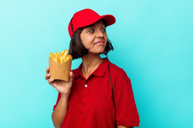 Młoda rasa mieszana kobieta pracownik restauracji fast food trzymający frytki na białym tle na niebieskim tle wygląda na uśmiechniętą, wesołą i przyjemną.