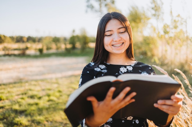 Młoda religijna chrześcijanka trzymająca Biblię i czytająca ją na zewnątrz w polu o zachodzie słońca.