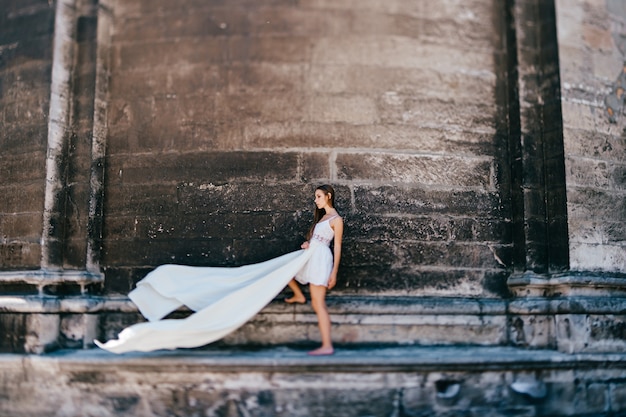 Młoda romantyczna elegancka dziewczyna w długiej białej sukni lejącej pozuje nad kamiennym murem