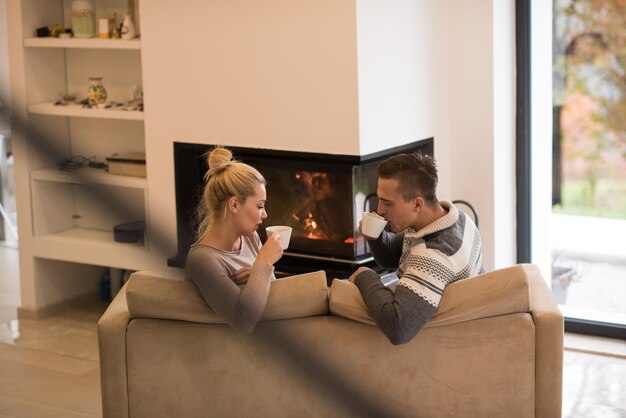 Młoda romantyczna para siedzi na kanapie przed kominkiem w domu, patrząc na siebie, rozmawiając i pijąc kawę w jesienny dzień