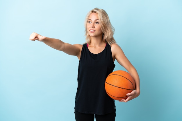 Młoda Rosjanka gra w koszykówkę na białym tle na niebieskiej ścianie, dając gest kciuki do góry