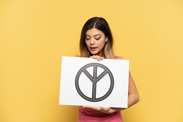 Młoda Rosjanka odizolowana na żółtym tle trzymająca afisz z symbolem pokoju