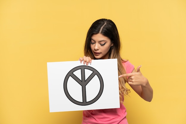 Młoda Rosjanka odizolowana na żółtym tle trzymająca plakat z symbolem pokoju i wskazująca go