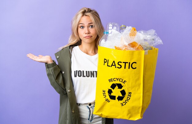 Młoda Rosjanka trzyma worek recyklingu pełen papieru do recyklingu na białym tle na fioletowej ścianie, mając wątpliwości podczas podnoszenia rąk