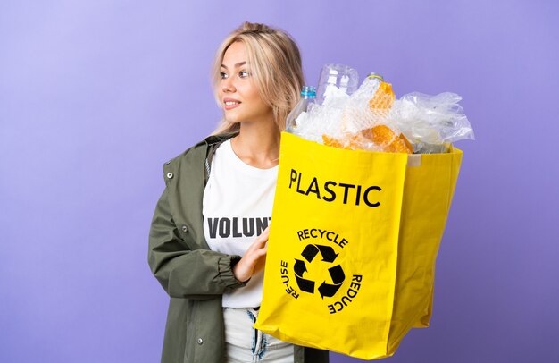 Młoda Rosjanka Trzyma Worek Recyklingu Pełen Papieru Do Recyklingu Na Białym Tle Na Fioletowej ścianie, Patrząc Z Boku I Uśmiechnięty