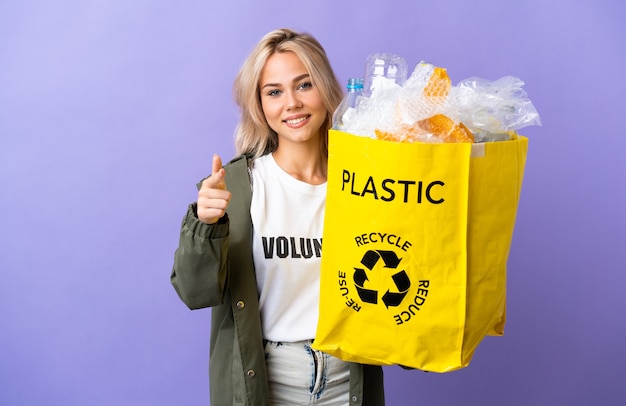 Młoda Rosjanka trzyma worek recyklingu pełen papieru do recyklingu na białym tle na fioletowej ścianie, wskazując na przód i uśmiechnięty