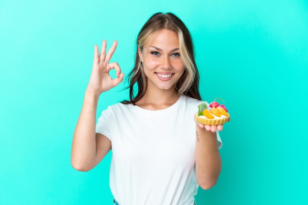 Młoda Rosjanka trzymająca słodki owoc na białym tle na niebieskim tle pokazująca znak ok palcami