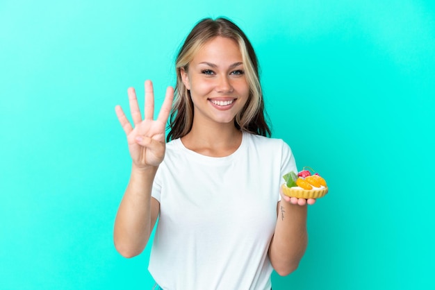 Młoda Rosjanka trzymająca słodki owoc na białym tle na niebieskim tle szczęśliwa i licząca cztery palcami