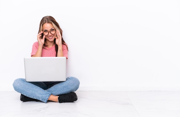 Młoda Rosjanka z laptopem siedzi na podłodze na białym tle na białym tle w okularach i zdziwiona