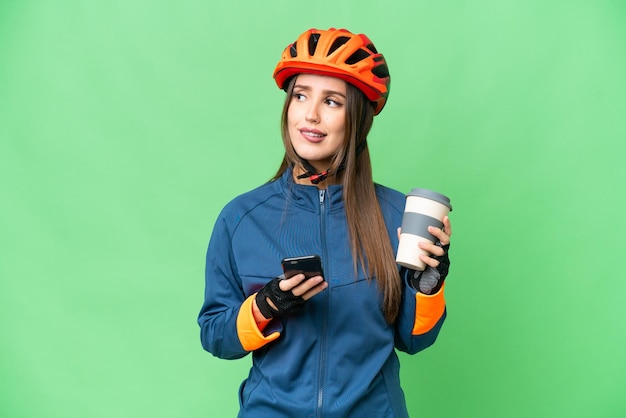Młoda rowerzystka na odizolowanym tle z kluczem chromatycznym, trzymając kawę na wynos i telefon komórkowy, myśląc o czymś