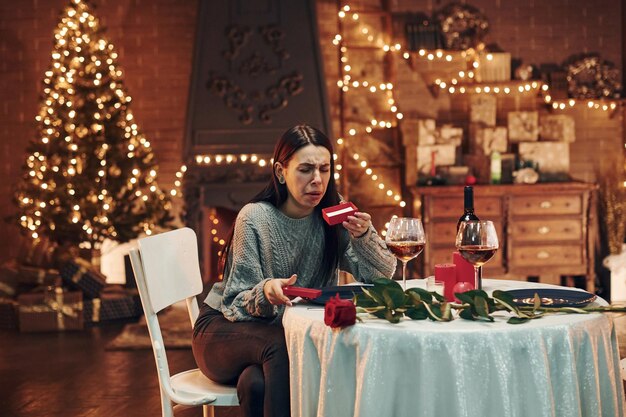 Młoda rozczarowana brunetka siedzi przy stole w restauracji i trzyma czerwone pudełko z prezentem