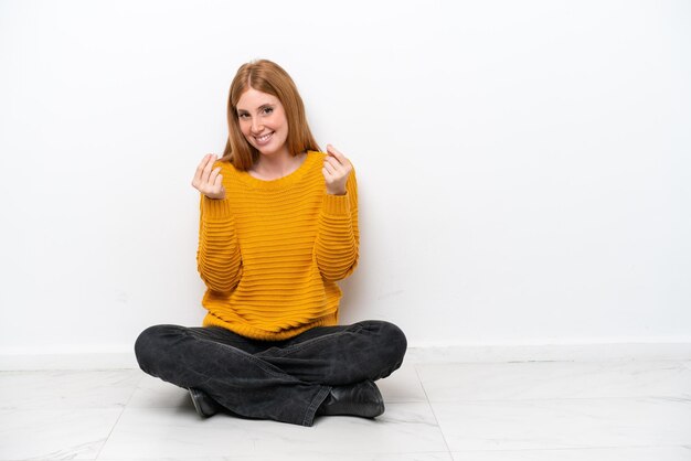 Zdjęcie młoda ruda kobieta siedzi na podłodze na białym tle robiąc gest pieniędzy