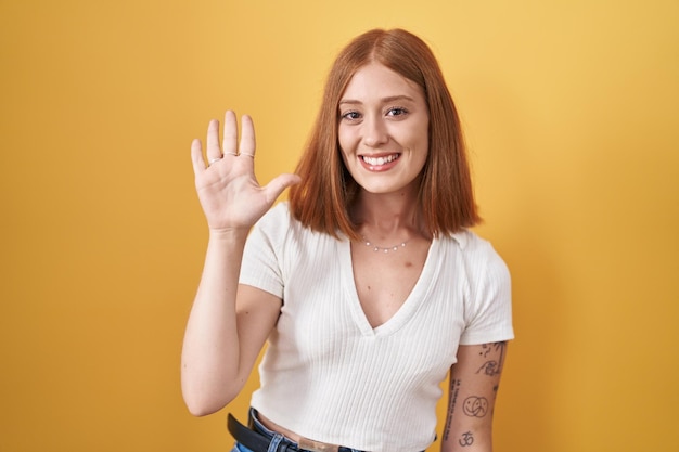 Młoda ruda kobieta stojąca na żółtym tle, pokazująca i wskazująca palcami numer pięć, uśmiechając się pewna siebie i szczęśliwa.