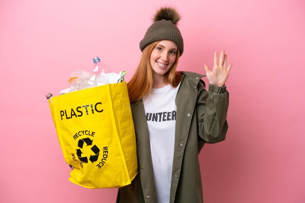 Młoda ruda kobieta trzyma torbę pełną plastikowych butelek do recyklingu na białym tle na różowym tle licząc pięć palcami