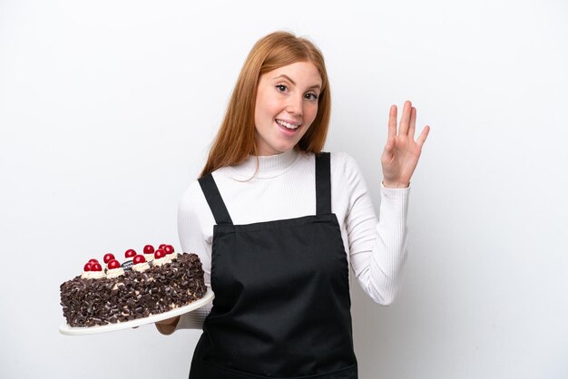 Młoda ruda kobieta trzyma tort urodzinowy na białym tle pozdrawiając ręką z szczęśliwym wyrazem twarzy