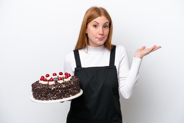 Młoda ruda kobieta trzymająca tort urodzinowy odizolowana na białym tle mająca wątpliwości podczas podnoszenia rąk