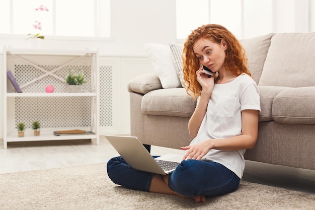 Młoda ruda kobieta wielozadaniowość. Siedząc na podłodze surfując po sieci i rozmawiając przez telefon komórkowy.