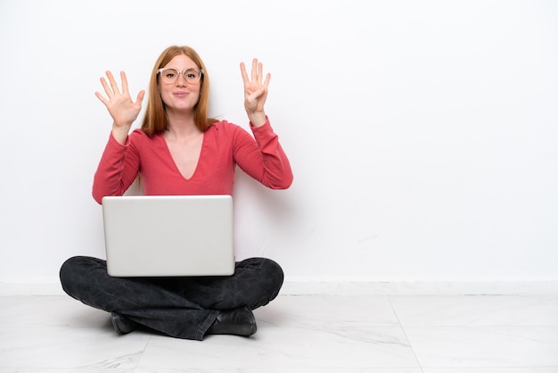 Młoda ruda kobieta z laptopem siedzi na podłodze na białym tle, licząc dziewięć palcami
