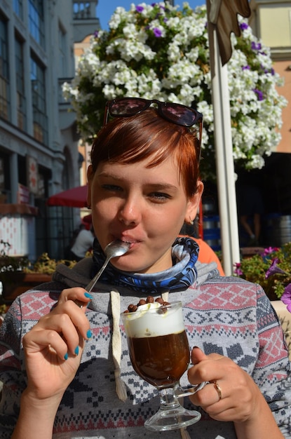 Młoda rudowłosa dziewczyna siedzi w cieniu w pobliżu kawiarni, pije cappuccino i liże łyżkę