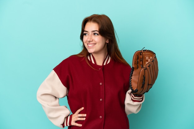 Młoda rudowłosa kobieta gra w baseball na białym tle na niebieskim tle pozuje z rękami na biodrach i uśmiecha się