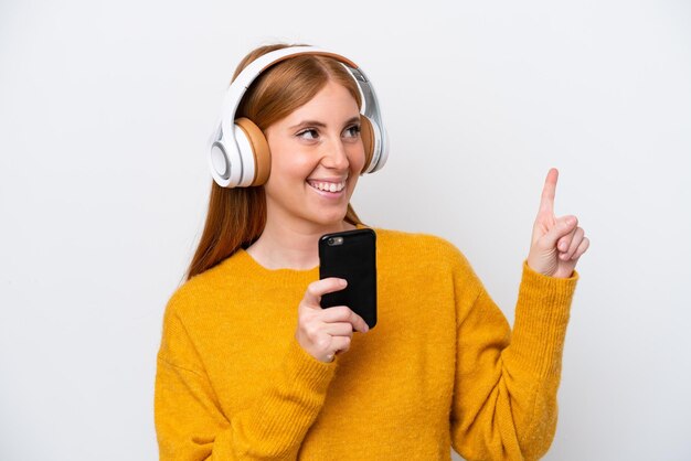 Młoda rudowłosa kobieta na białym tle słuchająca muzyki za pomocą telefonu komórkowego i śpiewania