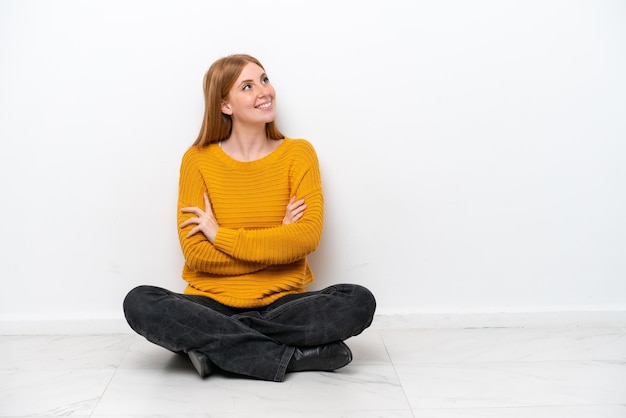 Młoda rudowłosa kobieta siedzi na podłodze na białym tle patrząc w górę podczas uśmiechania się