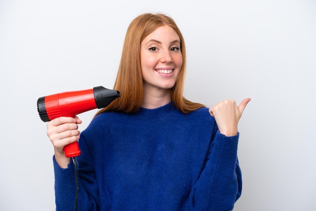 Młoda rudowłosa kobieta trzyma suszarkę na białym tle na białym tle, wskazując na bok, aby zaprezentować produkt