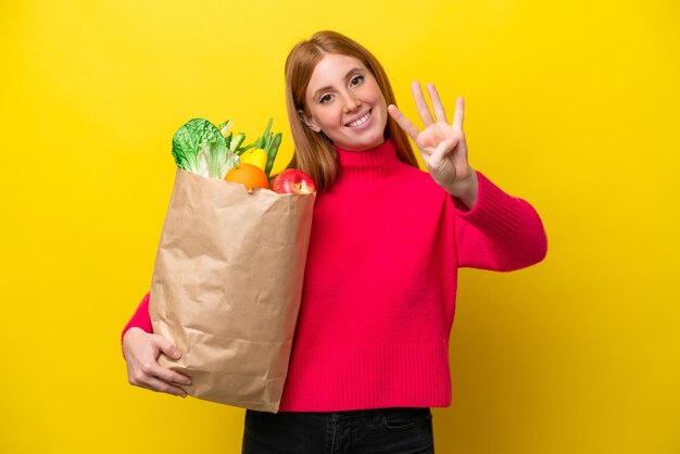 Młoda rudowłosa kobieta trzymająca torbę na zakupy spożywcze na żółtym tle szczęśliwa i licząca cztery palcami
