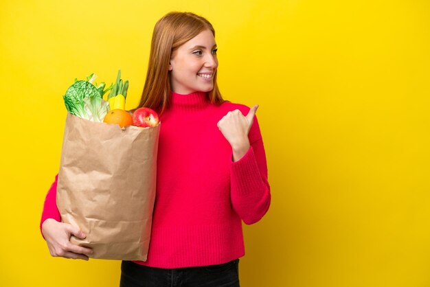 Młoda rudowłosa kobieta trzymająca torbę na zakupy spożywcze odizolowaną na żółtym tle wskazującą na bok, aby zaprezentować produkt