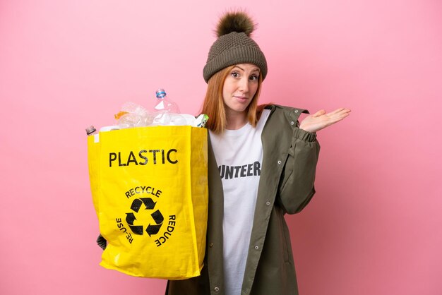 Młoda rudowłosa kobieta trzymająca torbę pełną plastikowych butelek do recyklingu na białym tle na różowym tle, mająca wątpliwości podczas podnoszenia rąk
