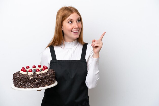Młoda rudowłosa kobieta trzymająca tort urodzinowy na białym tle zamierzająca zrealizować rozwiązanie, podnosząc palec w górę