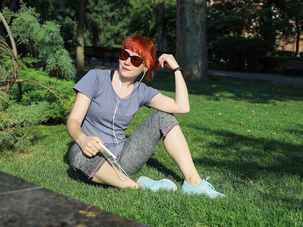 Młoda rudowłosa kobieta w stroju sportowym siedzi samotnie na trawie w parku słucha przez słuchawki