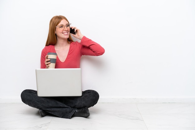 Młoda rudowłosa kobieta z laptopem siedząca na podłodze na białym tle trzymająca kawę na wynos i telefon komórkowy