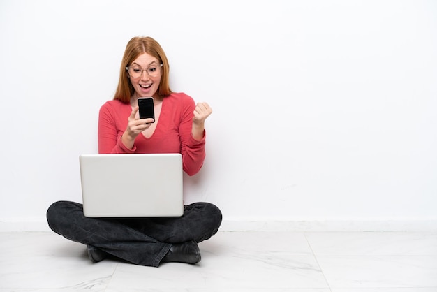 Młoda rudowłosa kobieta z laptopem siedząca na podłodze na białym tle zaskoczona i wysyłająca wiadomość