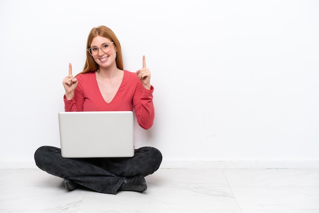 Młoda rudowłosa kobieta z laptopem siedzi na podłodze na białym tle i wskazuje na świetny pomysł