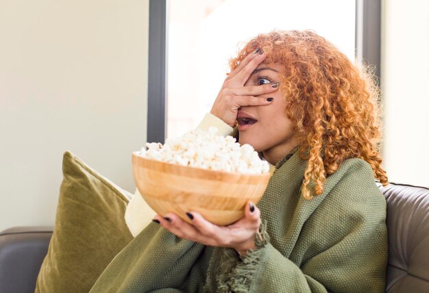 Młoda rudowłosa latynoska ładna kobieta jedząca popcorn na kanapie i oglądająca film