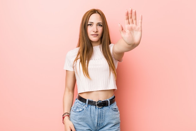 Młoda rudzielec imbirowa kobieta przeciw różowej ściennej pozyci z wyciągniętą ręką pokazuje znak stop, zapobieganie ci.