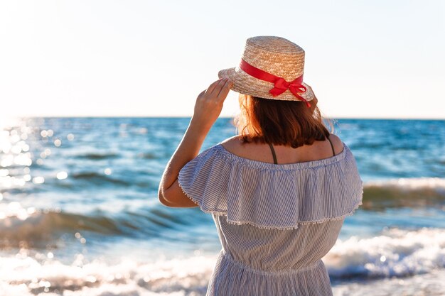 Młoda Samotna Kaukaska Rudowłosa Dziewczyna W Słomkowym Kapeluszu I Letniej Sukience Z Powrotem Na Plaży W Pobliżu Morza I Wygląda