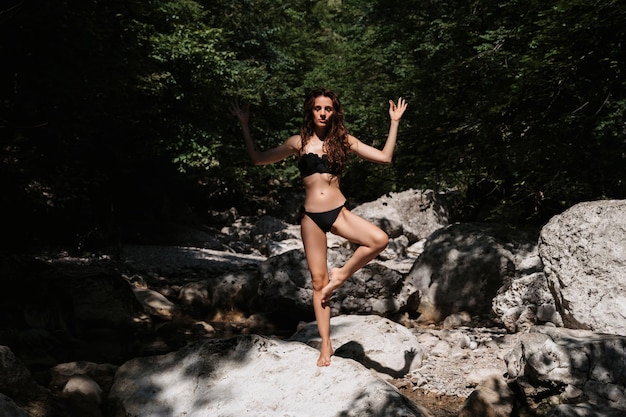 Młoda seksowna brunetka w czarnym stroju kąpielowym stoi w pozie jogi na skałach nad rzeką w naturze latem