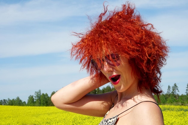 Młoda seksowna kaukaska kobieta z rudymi włosami i okularami przeciwsłonecznymi na żółtym tle pola rzepaku, letni dzień