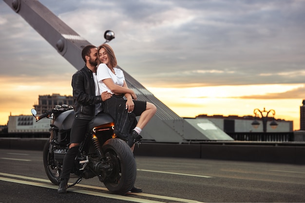 Młoda seksowna kobieta przytulanie ładny mężczyzna w stylowej czarnej skórzanej kurtce, siedząc na motocyklu sportowym na moście w mieście na zachód słońca i całując.
