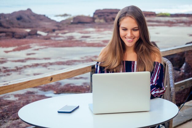 Młoda Seksowna Kobieta W Stylowym Kombinezonie W Paski Za Pomocą Laptopa I Telefonu Freelancer Na Tropikalnej Plaży Raju. Dziewczyna Freelancer Pracuje Siedząc W Letniej Kawiarni Praca Zdalna Na Tle Oceanu Indyjskiego