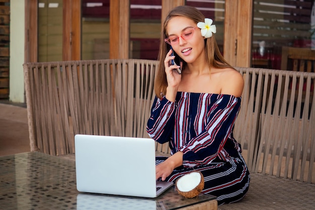 Młoda seksowna kobieta w stylowym kombinezonie w paski za pomocą laptopa i telefonu freelancer na tropikalnej plaży raju. Dziewczyna freelancer pracuje siedząc w letniej kawiarni praca zdalna na tle oceanu indyjskiego