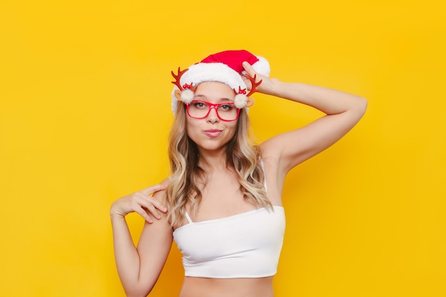 Młoda seksowna kobieta w świątecznych okularach z rogami jelenia na białym tle na żółtym tle