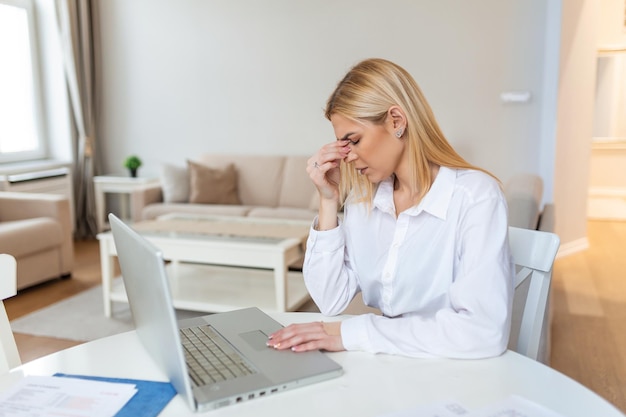Młoda sfrustrowana kobieta pracująca przy biurku przed laptopem cierpiąca na przewlekłe codzienne leczenie bólu głowy online umawianie się na konsultację medyczną promieniowanie elektromagnetyczne wynagrodzenie chorobowe