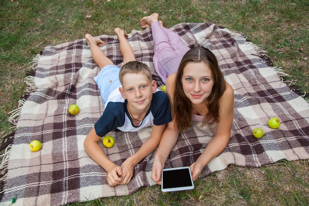 Zdjęcie młoda siostra i brat z piegami na twarzach, leżąc na pled i za pomocą tabletu w parku. widok z góry, patrząc na aparat z uśmiechem.