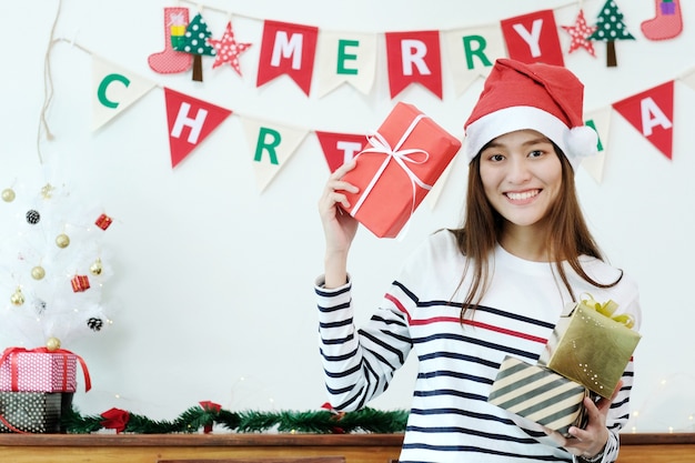Młoda śliczna azjatykcia kobieta jest ubranym Santa kapelusz uśmiecha się Bożenarodzeniowych prezentów pudełka i trzyma.