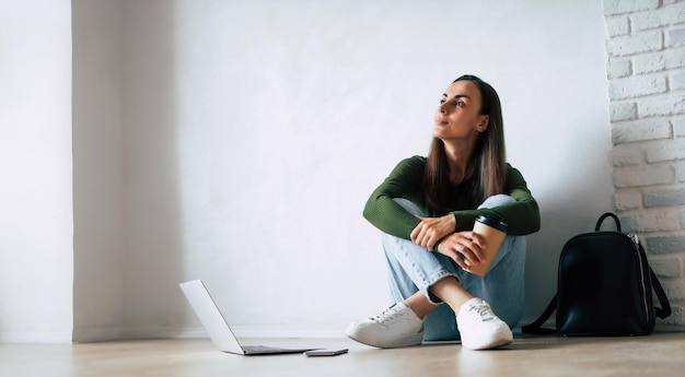 Młoda, śliczna, pewna siebie studentka w luźnych ubraniach używa laptopa i uśmiecha się, siedząc na podłodze na szarym tle Studia koncepcja absolwenta uczelni wyższej